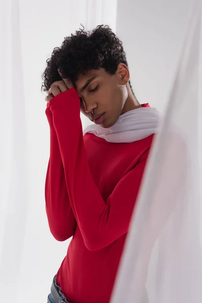 African american man in red turtleneck posing near white tulle cloth with closed eyes and hands near face - foto de stock