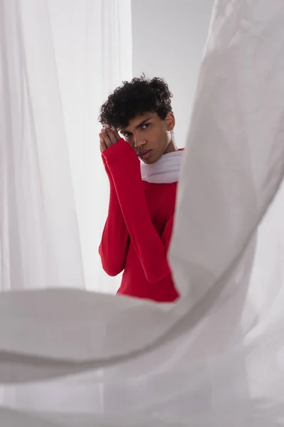 Stylish african american man in red turtleneck posing with praying hands near white blurred drapery — Fotografia de Stock