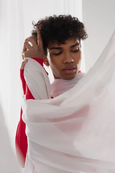 Pierced african american man with trendy hairstyle posing near white drapery - foto de stock