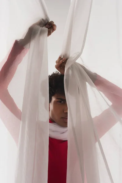 African american man looking at camera while hiding behind white transparent drapery — Fotografia de Stock