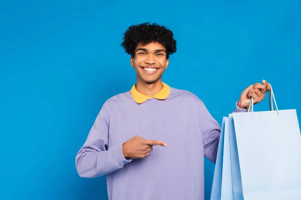 Fröhlicher afrikanisch-amerikanischer Mann zeigt vereinzelt auf blaue Einkaufstüten — Stockfoto