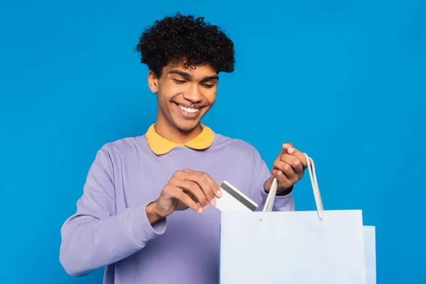 Joyful african american man putting credit card into shopping bag isolated on blue — Photo de stock