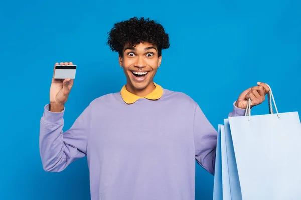 Erstaunt afrikanisch-amerikanischer Mann hält Einkaufstaschen und Kreditkarte isoliert auf blauem Grund — Stockfoto