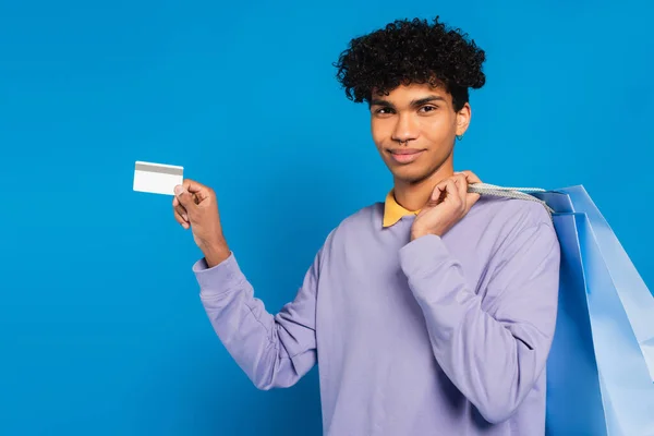 Young african american man with shopping bags showing credit card isolated on blue — Photo de stock