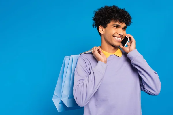Happy african american man with shopping bags talking on mobile phone isolated on blue - foto de stock