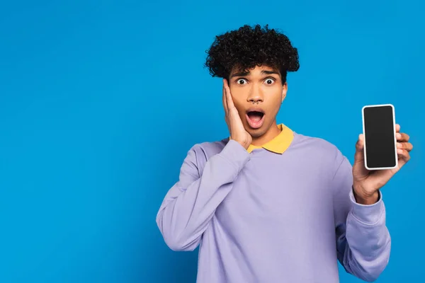 Shocked african american man touching face while showing cellphone with blank screen isolated on blue - foto de stock