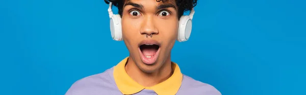 Amazed african american man in headphones looking at camera isolated on blue, banner — Foto stock