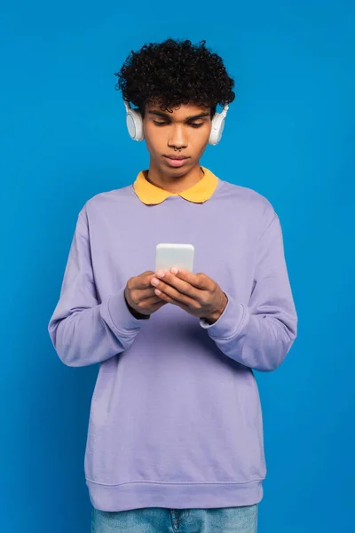 Молодий афроамериканський чоловік в навушниках, використовуючи мобільний телефон ізольований на синьому — стокове фото