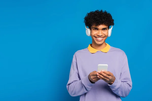 Joyful african american man with smartphone listening music in headphones isolated on blue — Fotografia de Stock