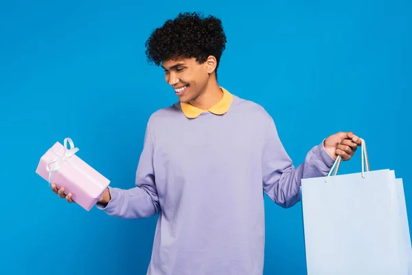 Fröhlicher afrikanisch-amerikanischer Mann in fliederfarbenem Pullover mit Einkaufstaschen und Geschenk isoliert auf blauem Grund — Stockfoto