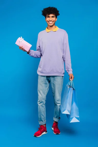 Volle Länge Ansicht des afrikanisch-amerikanischen Mannes, der mit Einkaufstaschen steht und auf blauem Hintergrund präsent ist — Stockfoto