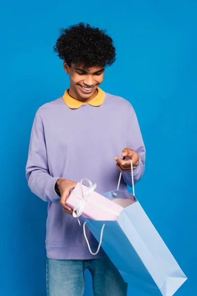 Веселий афроамериканський чоловік вставляє подарунок у паперовий мішок ізольовано на синьому — стокове фото