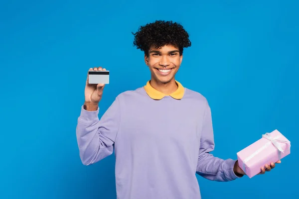 Happy african american man with credit card and gift box looking at camera isolated on blue — Foto stock