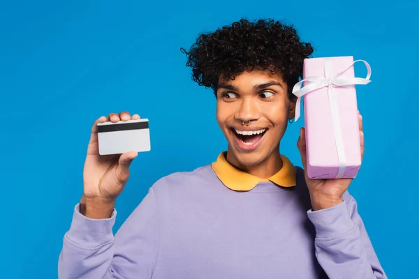 Excited african american man showing credit card and gift box isolated on blue — Foto stock