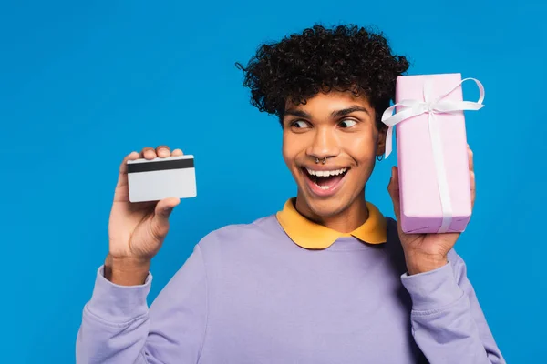 Excited african american man showing credit card and gift box isolated on blue - foto de stock