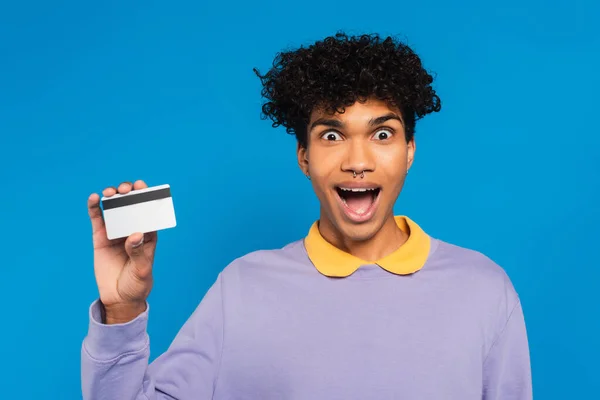 Astonished african american man in violet pullover showing credit card isolated on blue — Foto stock