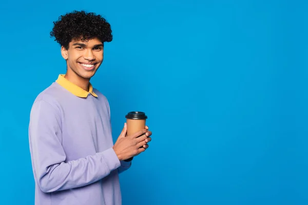 Веселый африканский американец в фиолетовом свитере с желтым воротником, держащий бумажную чашку, изолированную на голубом — стоковое фото