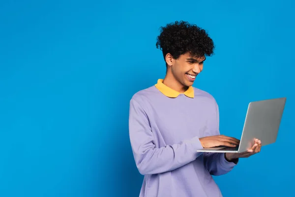 Happy african american man in violet pullover using laptop isolated on blue — Fotografia de Stock