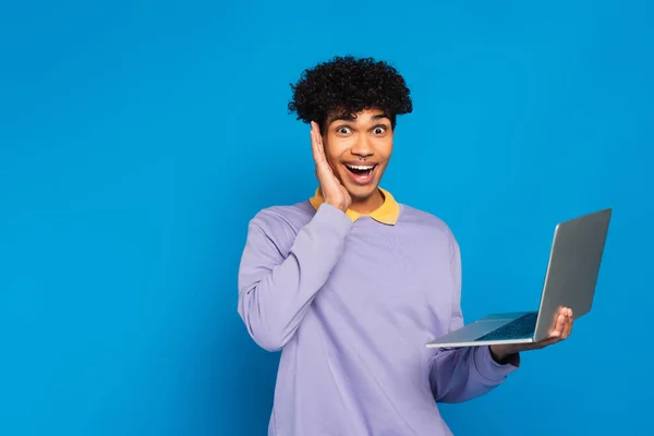 Amazed african american student with laptop touching face while looking at camera isolated on blue — Fotografia de Stock