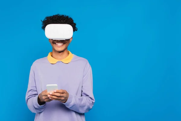 Веселый африканский американец с помощью смартфона в VR гарнитуре изолированы на синий — стоковое фото