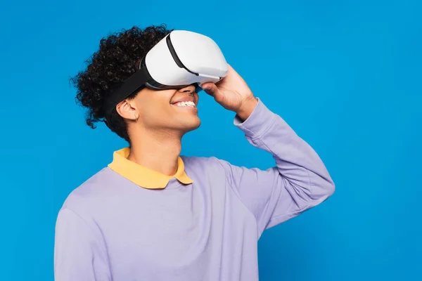 Joyful african american man in purple jumper and vr headset looking away isolated on blue - foto de stock