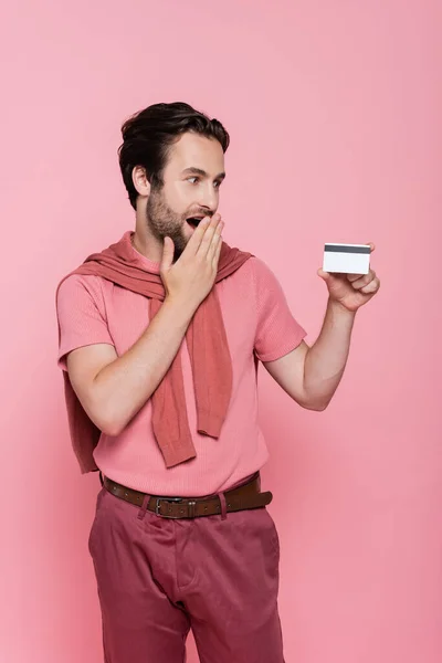 Uomo stupito guardando la carta di credito isolata su rosa — Foto stock