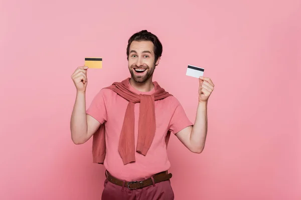 Позитивний чоловік тримає кредитні картки і дивиться на камеру ізольовано на рожевому — стокове фото