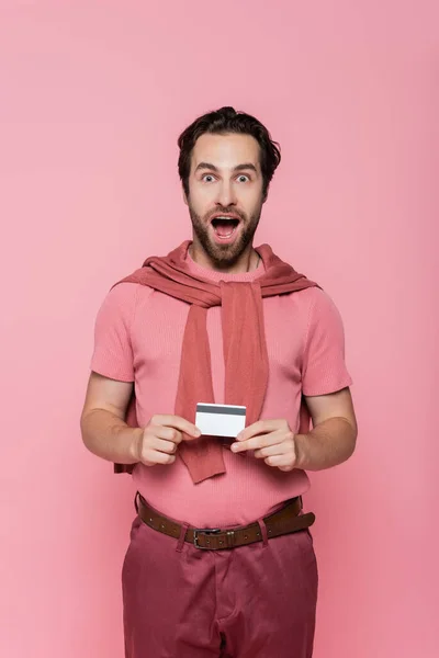 Hombre sorprendido con tarjeta de crédito mirando la cámara aislada en rosa - foto de stock