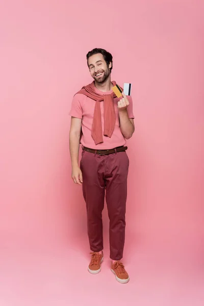 Hombre alegre sosteniendo tarjetas de crédito y mirando a la cámara en el fondo rosa - foto de stock