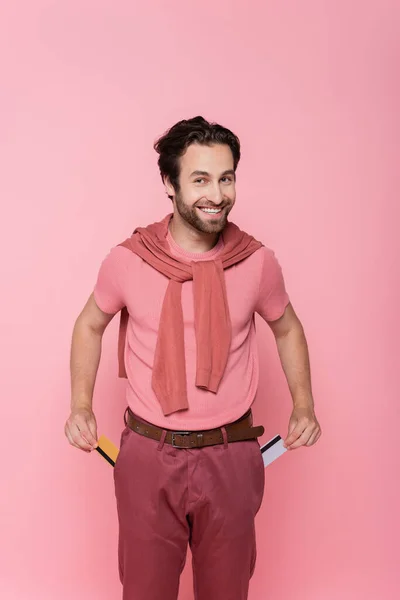 Hombre sonriente sosteniendo tarjetas de crédito cerca de bolsillos de pantalones aislados en rosa - foto de stock