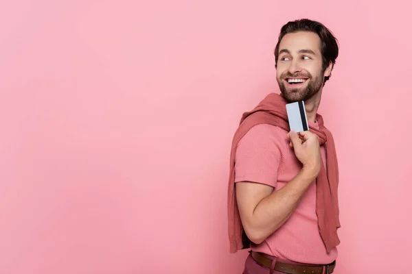 Bruna uomo sorridente mentre tiene la carta di credito e distogliendo lo sguardo isolato sul rosa — Foto stock