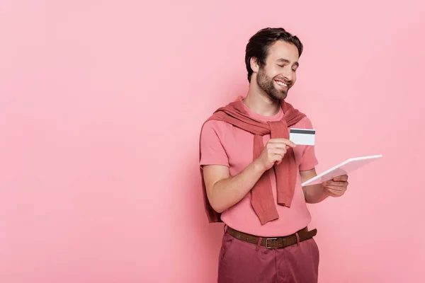 Улыбающийся мужчина держит цифровой планшет и кредитную карту изолированы на розовый — стоковое фото