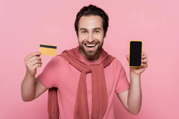 Sonriente hombre sosteniendo teléfono inteligente y tarjeta de crédito aislado en rosa - foto de stock