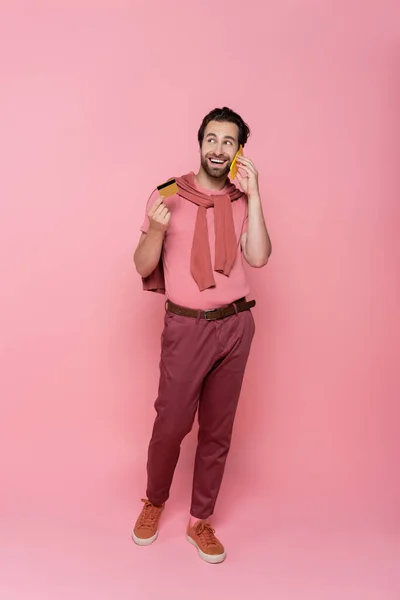 Smiling man talking on smartphone and holding credit card during online shopping on pink background — Stock Photo