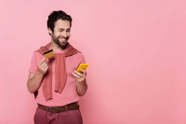 Positive man holding credit card and using cellphone isolated on pink — Stock Photo