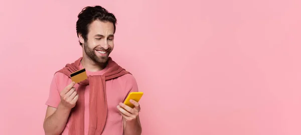 Sonriente hombre mirando el teléfono inteligente y la celebración de la tarjeta de crédito durante e compras aislado en rosa, pancarta - foto de stock