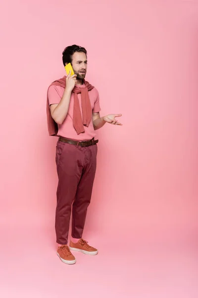 Longitud completa del hombre confundido hablando en el teléfono móvil sobre fondo rosa - foto de stock