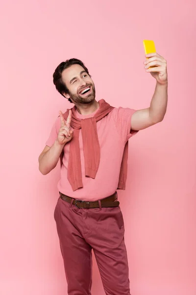 Uomo sorridente che mostra il gesto della vittoria mentre prende selfie su smartphone isolato su rosa — Foto stock