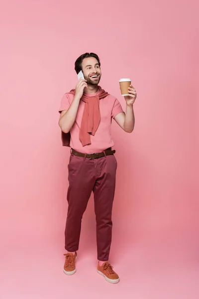 Pleine longueur de sourire homme parlant sur smartphone et tenant tasse en papier sur fond rose — Photo de stock