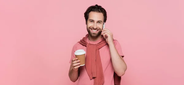 Young man talking on mobile phone and holding takeaway drink isolated on pink, banner — Stock Photo