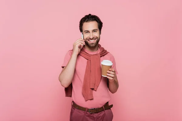 Positiver Mann blickt in die Kamera, während er mit dem Handy telefoniert und Kaffee to go auf rosa hält — Stockfoto