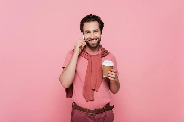 Homme positif regardant la caméra tout en parlant sur un téléphone portable et en tenant du café pour aller isolé sur rose — Photo de stock