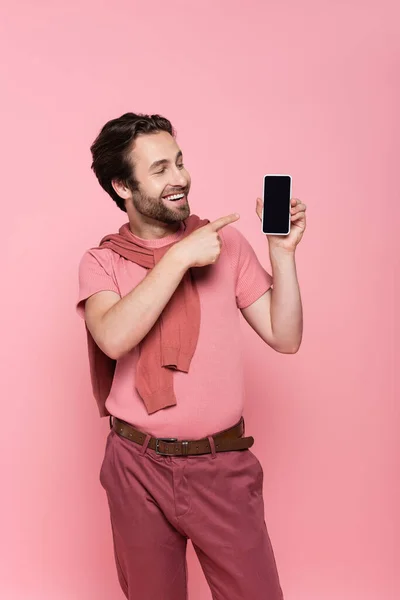Улыбающийся мужчина указывает на смартфон с пустым экраном, изолированным на розовом — стоковое фото
