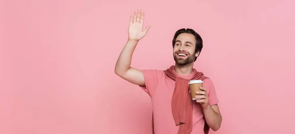 Homem positivo segurando copo de papel e mão acenando isolado em rosa, banner — Fotografia de Stock