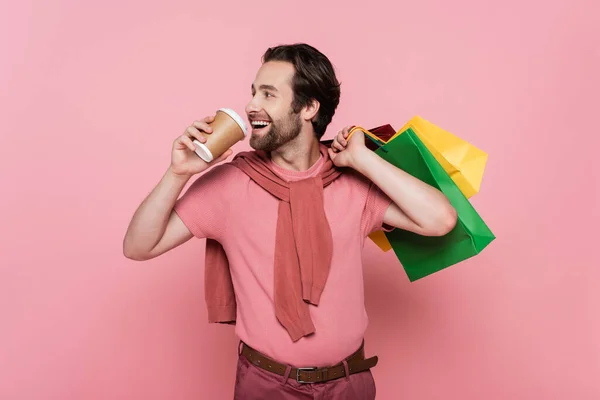 Vista lateral de un joven feliz sosteniendo bolsas de compras y taza de papel sobre fondo rosa - foto de stock