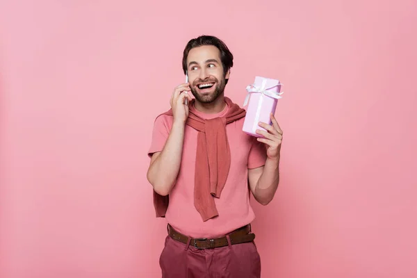 Aufgeregter Mann telefoniert mit Handy und hält Geschenk isoliert auf rosa — Stockfoto