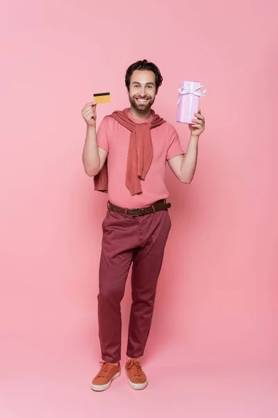 Улыбающийся мужчина с кредиткой и подарочной коробкой на розовом фоне — стоковое фото