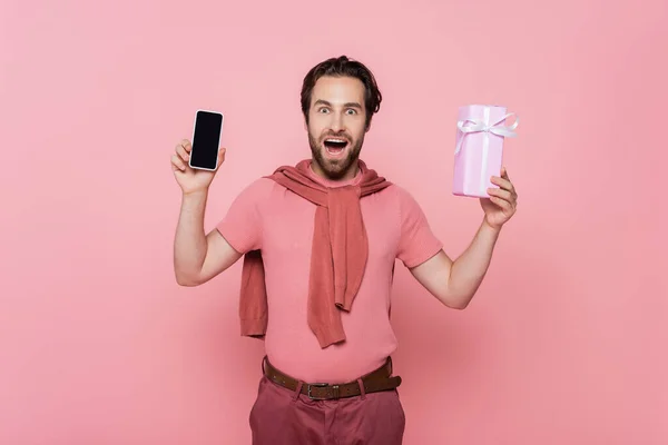 Astonished man holding smartphone with blank screen and present isolated on pink — Stock Photo