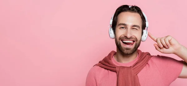 Homme souriant dans les écouteurs cligner des yeux et sortir la langue isolée sur rose, bannière — Photo de stock