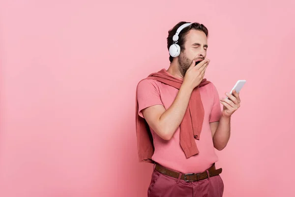 Молодий чоловік в навушниках позіхає, тримаючи смартфон ізольовано на рожевому — стокове фото